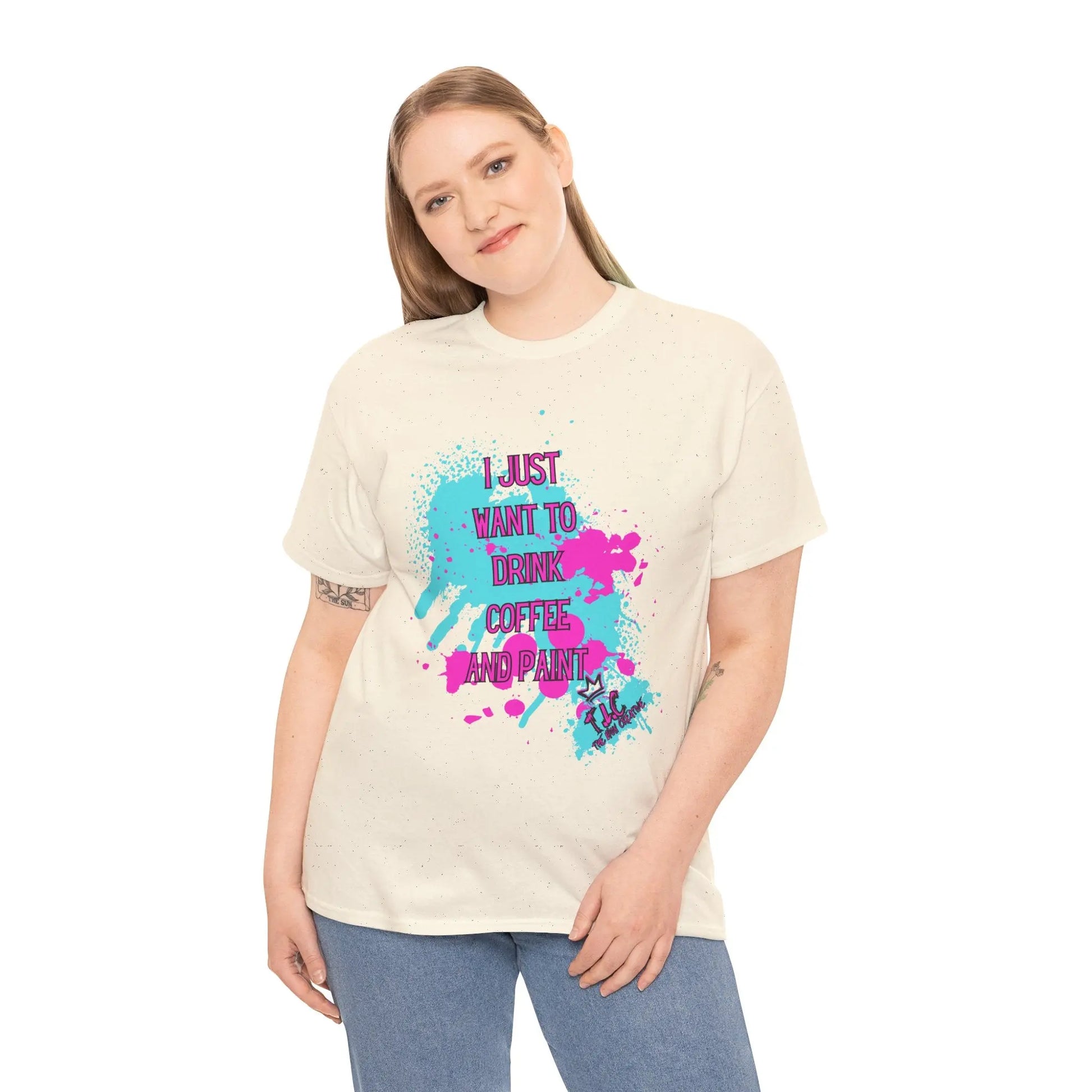 A person with long hair is wearing the "I Just want to Drink Coffee T-shirt," a cream-colored tee adorned with colorful splatters and the phrase "I just want to drink coffee and paint" in vibrant letters. Made from 100% cotton under the US Cotton Trust Protocol, this classic fit shirt perfectly complements their cheerful smile against a plain white background.