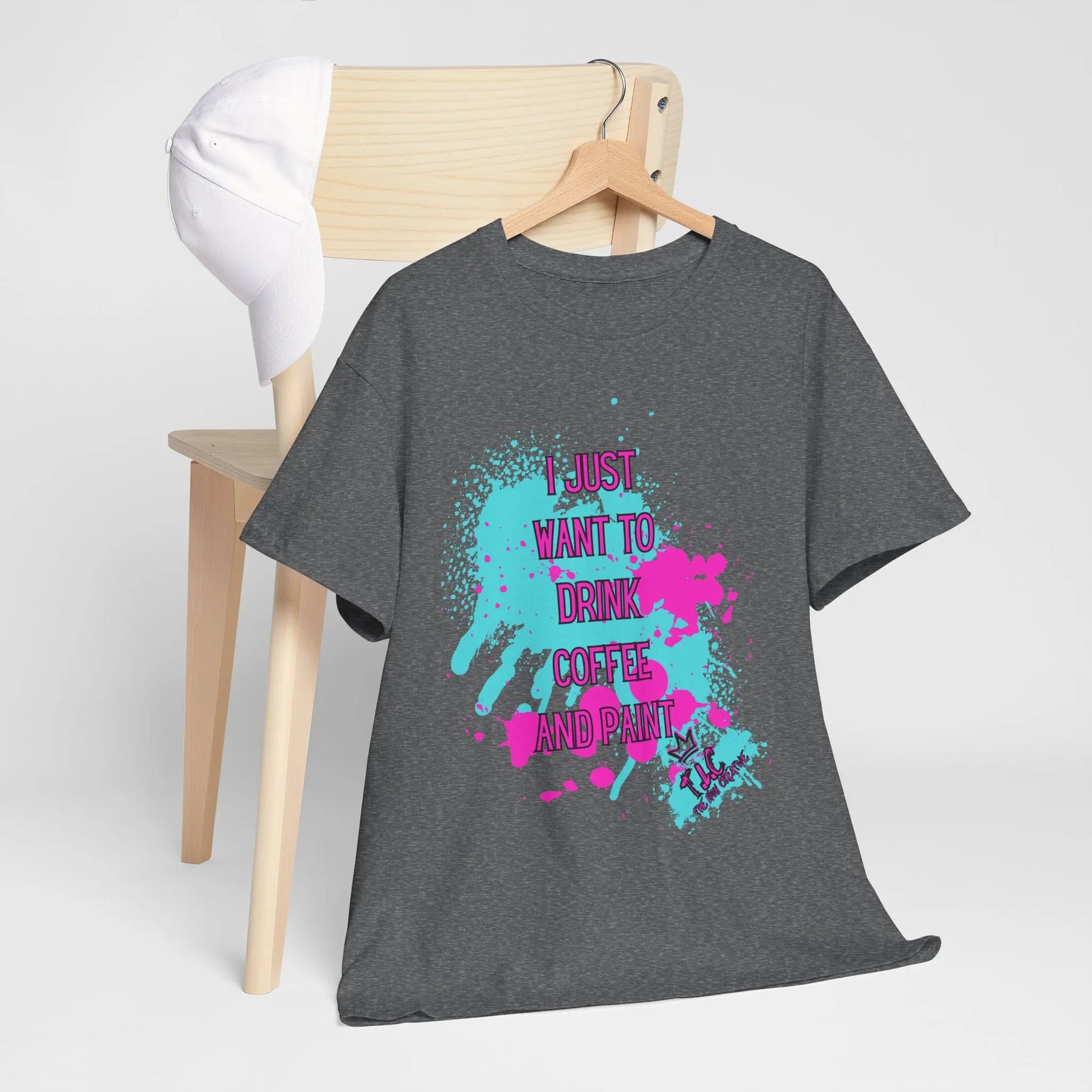 An ethically made gray cotton T-shirt, named "I Just Want to Drink Coffee," with colorful paint splatters and the words "I JUST WANT TO DRINK COFFEE AND PAINT," is draped over a wooden chair. A white cap rests on the chair's back, while the sustainable T-shirt is adorned with pink, blue, and purple accents.