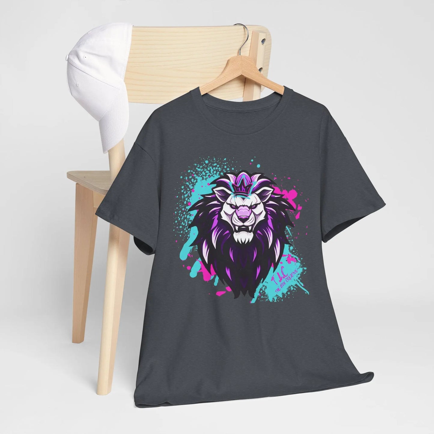 The Lyon T-shirt, in a classic fit and gray color, showcases a vibrant roaring lion graphic with purple and white highlights, accented by splashes of teal and pink paint. Crafted from 100% sustainable cotton, it elegantly drapes over a wooden chair, complemented by a white baseball cap resting on the backrest.