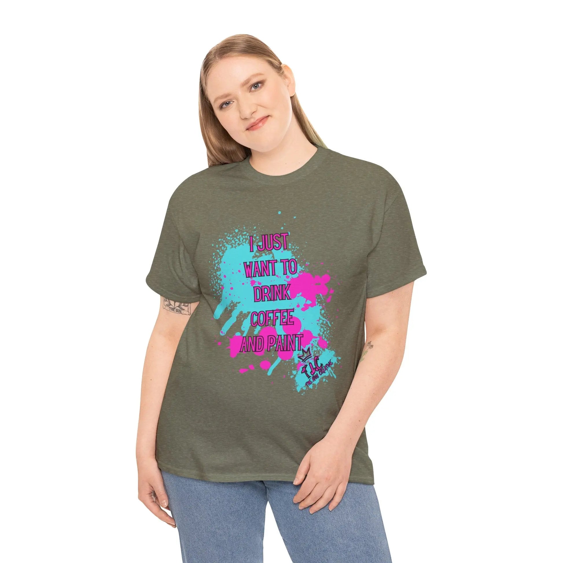 A person with long hair is wearing the "I Just Want to Drink Coffee T-shirt," a classic-fit, green cotton garment adorned with vibrant paint splashes and the phrase "I just want to drink coffee and paint." They are smiling against a plain white background, proudly supporting sustainable production.