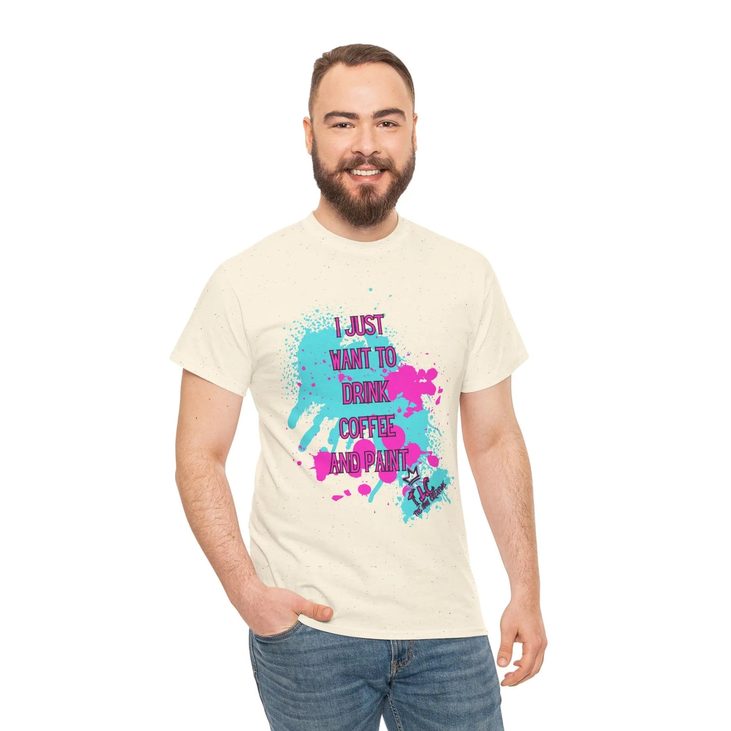 A bearded man is wearing the "I Just Want to Drink Coffee T-shirt," a beige, 100% cotton shirt with colorful splatters and the phrase "I JUST WANT TO DRINK COFFEE AND PAINT." Smiling, he stands with his right hand in his blue jeans pocket, highlighting its classic comfort fit.