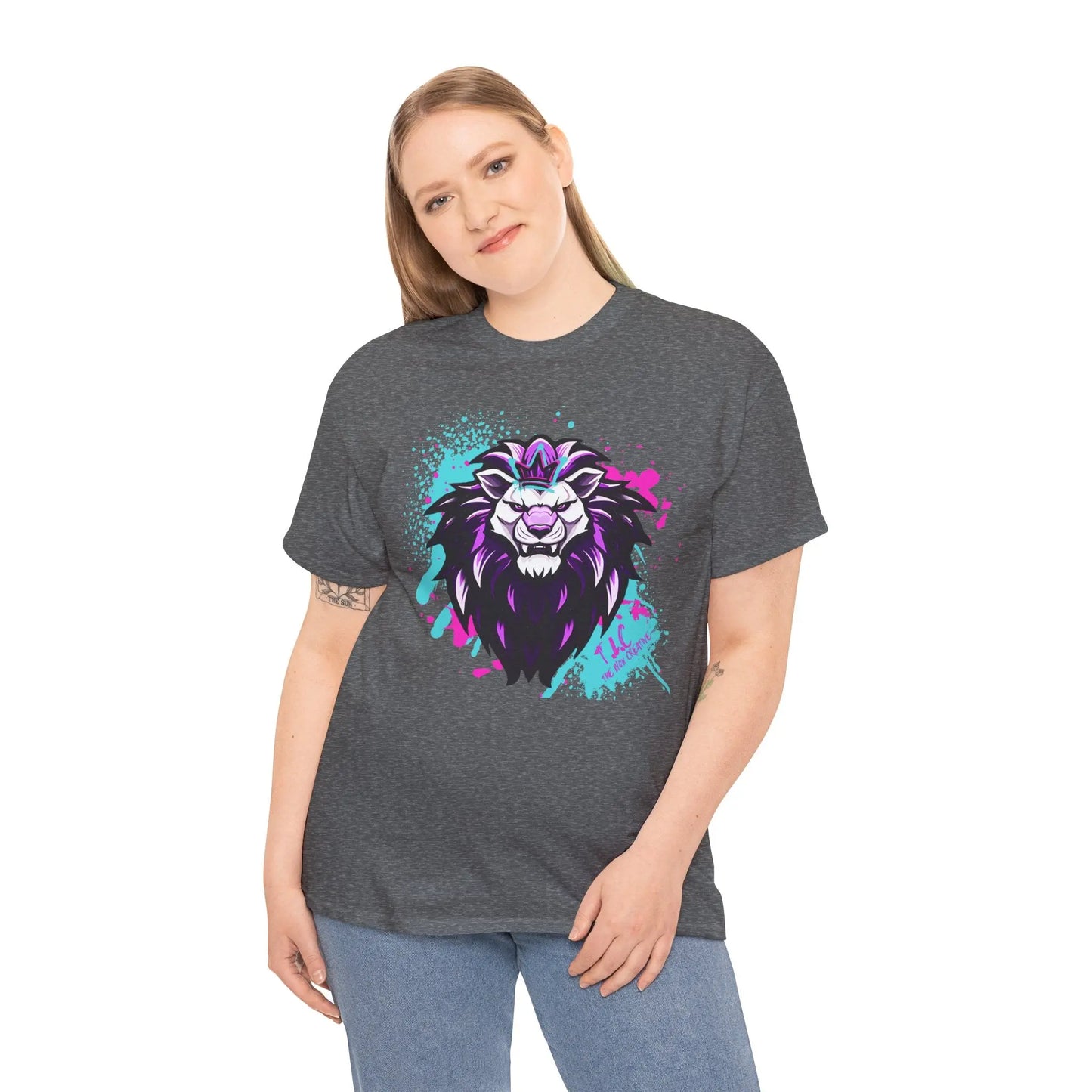 A person with long hair smiles while wearing The Lyon T-shirt, featuring a colorful lion design in purple, pink, and blue tones on a gray 100% cotton fabric. They stand against a plain white backdrop.