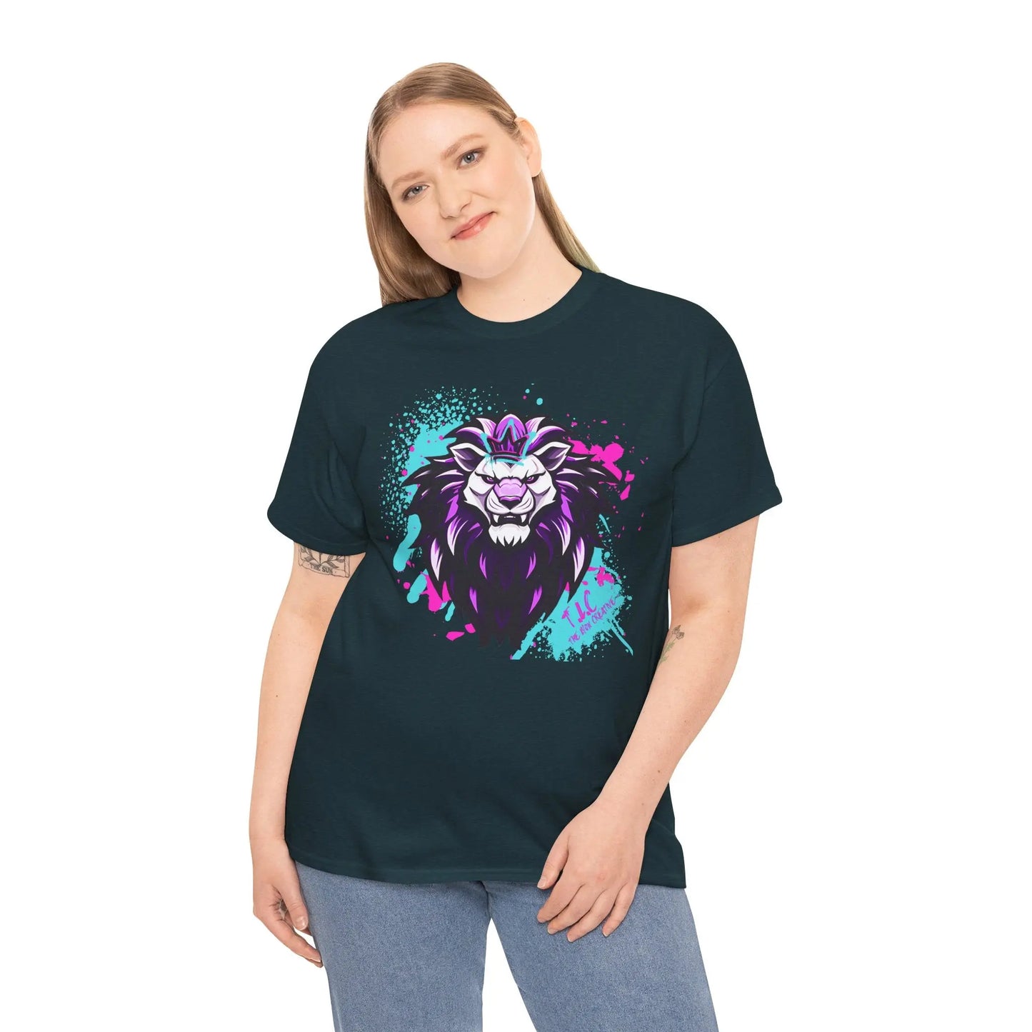 A person with long hair wearing The Lyon T-shirt, made from 100% cotton in dark green, showcasing a bold and colorful lion design in purple, white, and teal. They are also dressed in light blue jeans, standing against a white background.