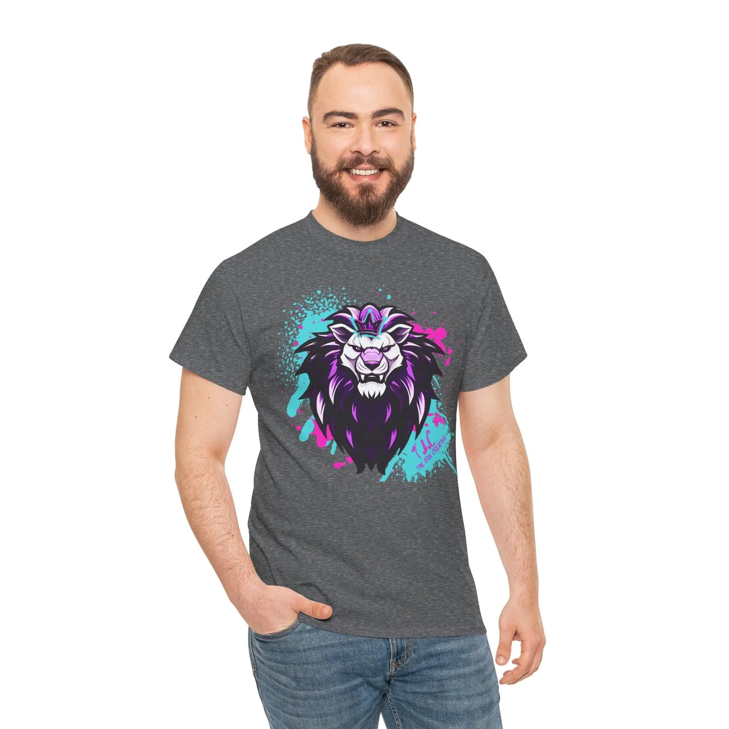 A man with a beard and short hair smiles while wearing The Lyon T-shirt, a gray 100% cotton shirt showcasing a vibrant lion graphic. The lion's mane is artistically illustrated in purple, white, and turquoise splashes. This classic fit shirt is sustainably produced to maintain quality as the man poses against a white background with one hand in his pocket.