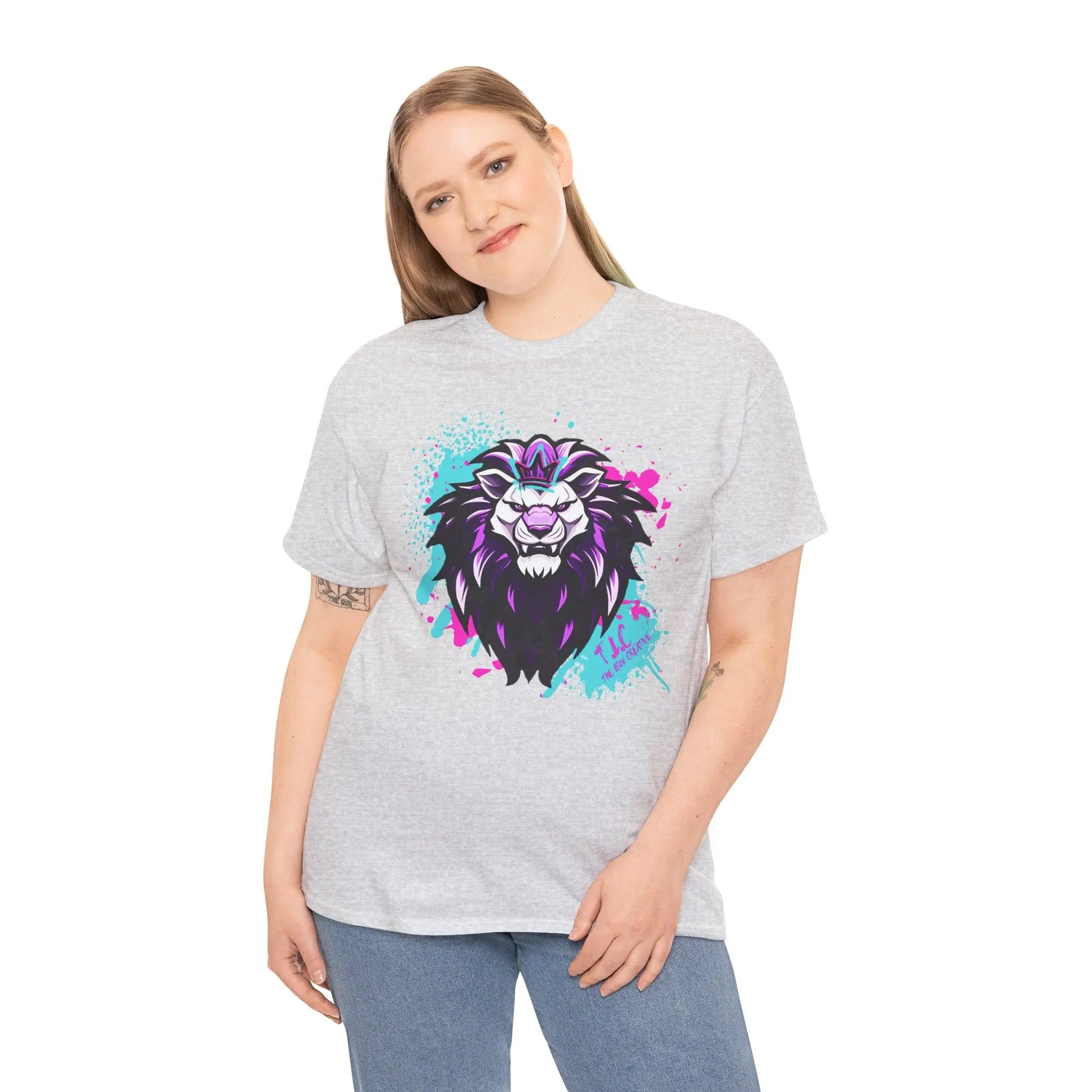 Wearing "The Lyon T-shirt," a person with long hair showcases a colorful and artistic lion design on a gray, 100% cotton fabric that offers a classic fit. They are standing against a plain white background, smiling slightly, and have one hand in their pocket.