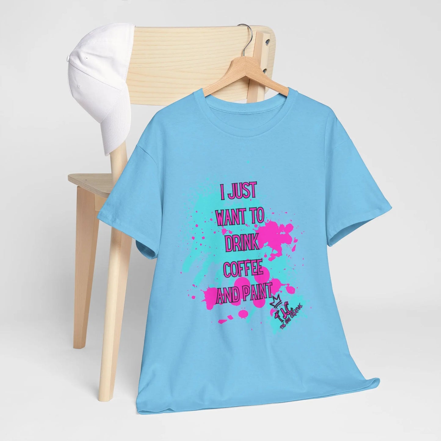 An "I Just Want to Drink Coffee T-shirt," featuring a blue cotton fabric with colorful paint splatters and the text "I just want to drink coffee and paint," is draped over a wooden chair. Designed for a classic fit, it complements the white cap resting on the chair's backrest.