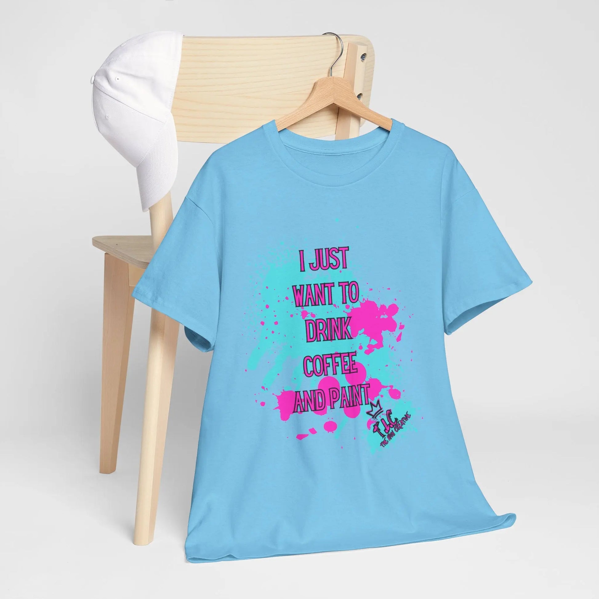 An "I Just Want to Drink Coffee T-shirt," featuring a blue cotton fabric with colorful paint splatters and the text "I just want to drink coffee and paint," is draped over a wooden chair. Designed for a classic fit, it complements the white cap resting on the chair's backrest.