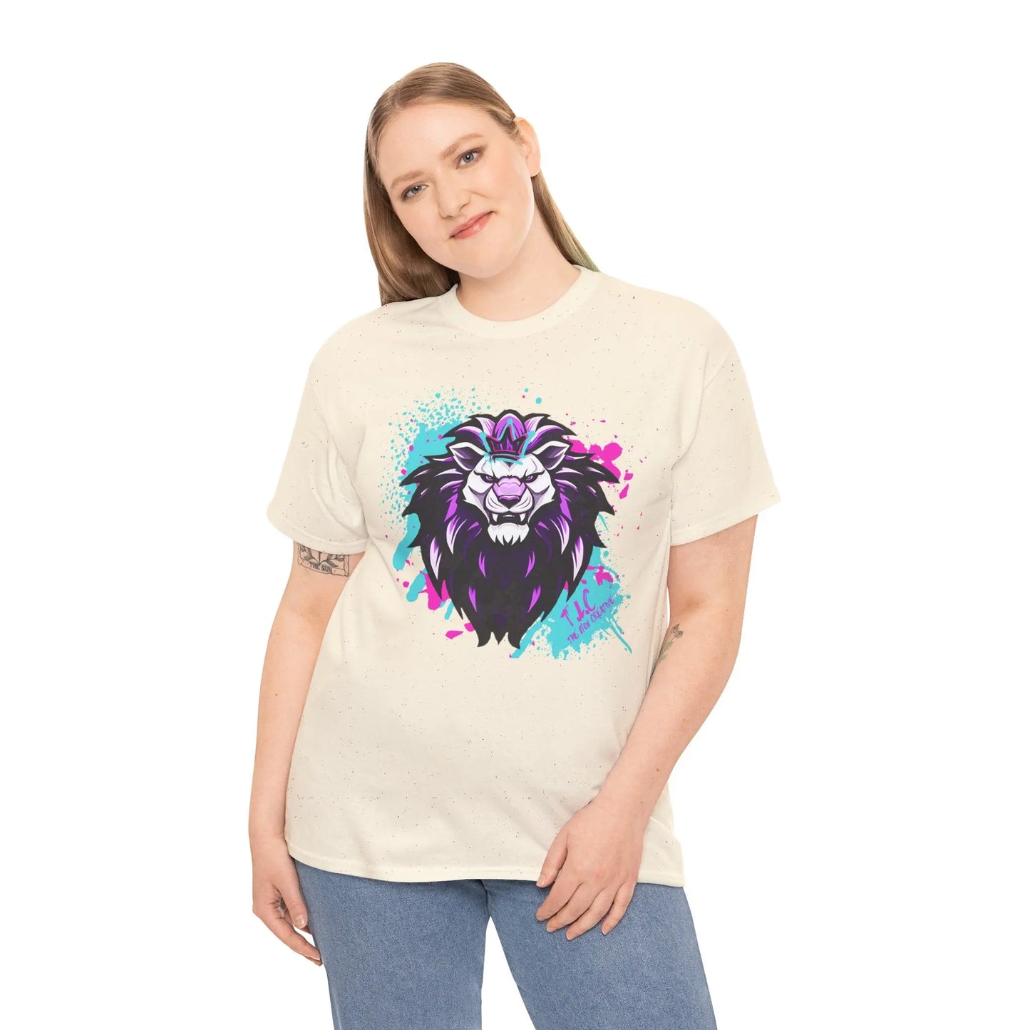 A person with long hair stands and smiles, wearing The Lyon T-shirt, which features a vibrant lion graphic adorned with purple and blue splashes. This classic fit, 100% cotton tee pairs perfectly with their blue jeans against the plain white background.