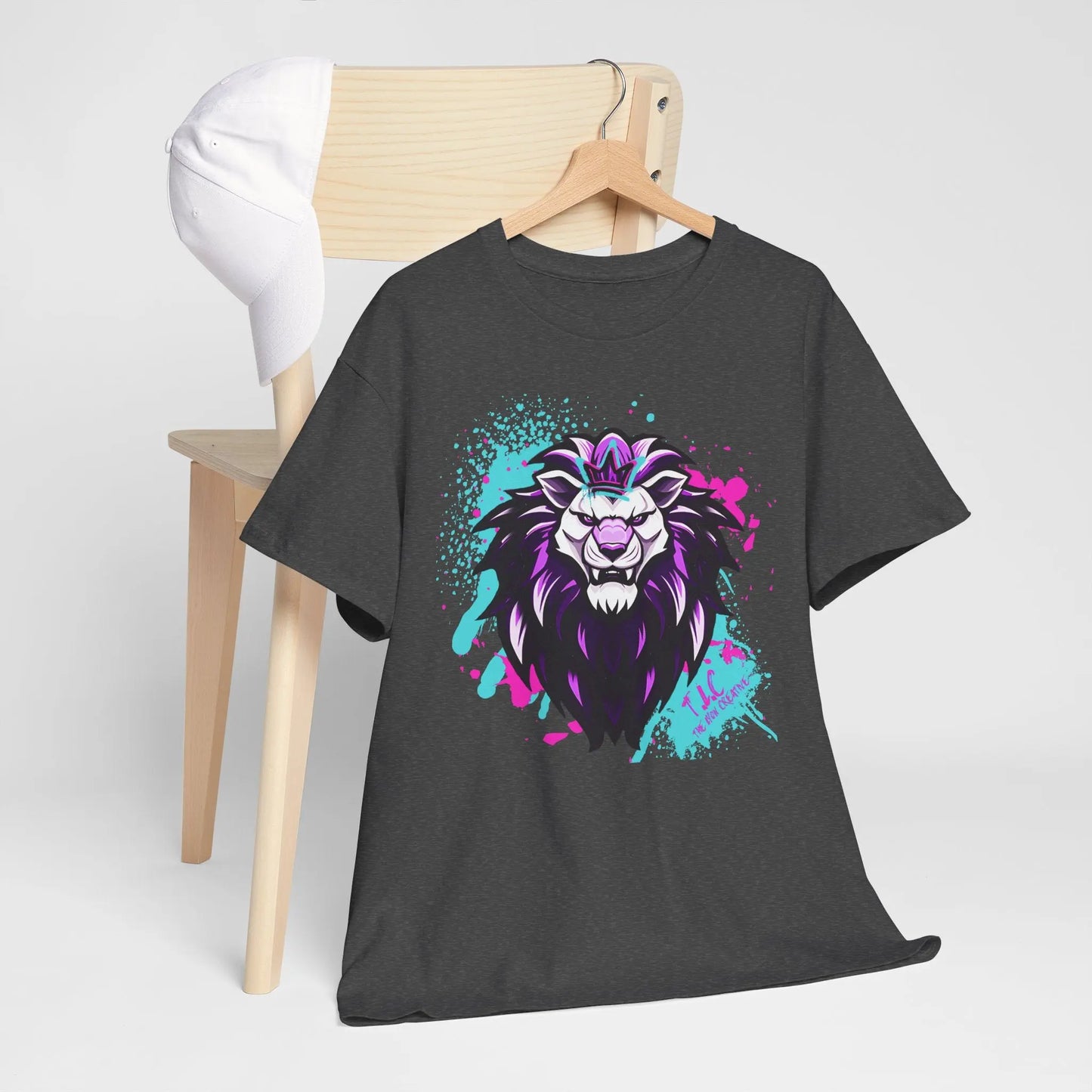 A dark Gildan shirt, named The Lyon T-shirt, showcases a vibrant and colorful graphic of a lion's head in purple, blue, and pink hues as it hangs on a wooden chair. Crafted from 100% cotton, its quality complements the white baseball cap resting on the backrest against a plain background.