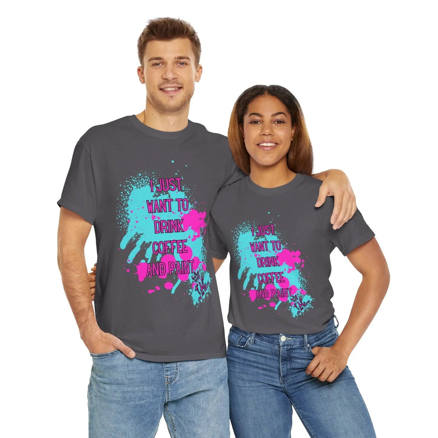 A man and a woman, smiling and standing together, each wear an "I Just want to Drink Coffee" T-shirt in classic-fit dark gray, featuring colorful paint splatters and the phrase "I just want to drink coffee and paint." These 100% cotton sustainable shirts are a perfect match for their creative spirits as they embrace one another.