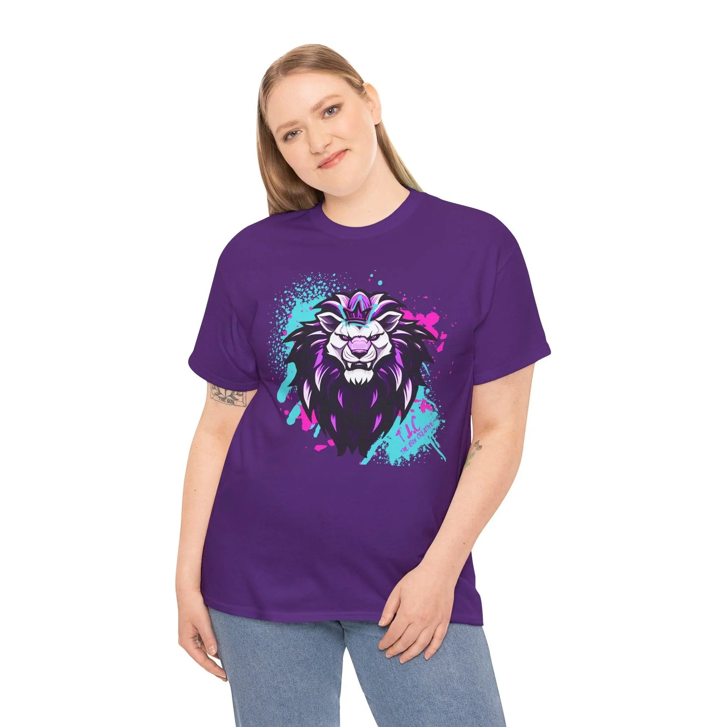 A person wearing The Lyon T-shirt, a classic fit purple top crafted from 100% cotton, showcases a striking graphic of a fierce lion's head with vivid purple and teal splashes. The person, with long hair, stands against a plain white background, highlighting the sustainably produced garment.