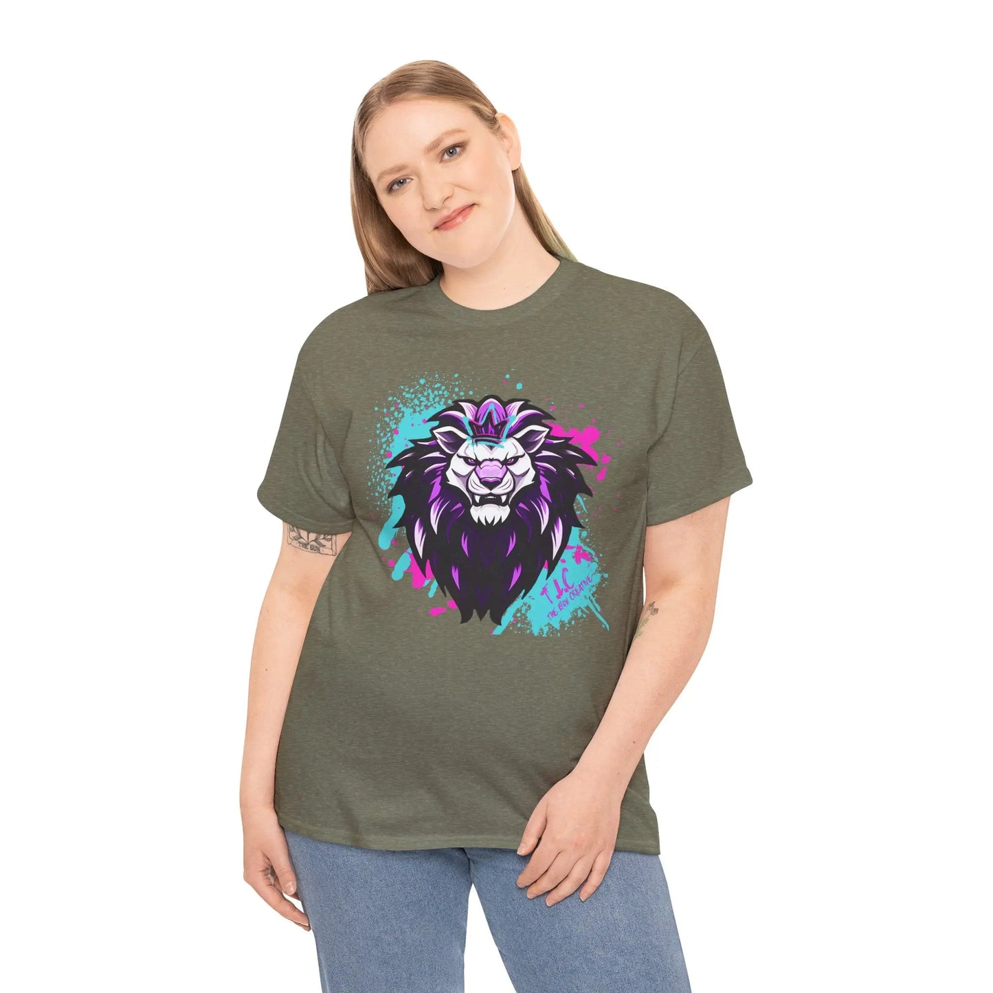 A person with long hair stands confidently, wearing The Lyon T-shirt, a classic fit olive green shirt made from 100% cotton. It showcases a vibrant, stylized lion design in purple and white with splashes of pink and teal. They also wear blue jeans and have one arm relaxed at their side.