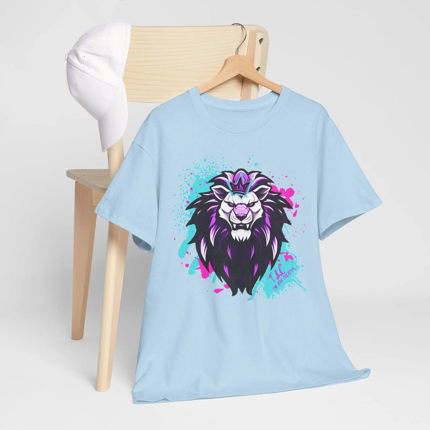 The Lyon T-shirt, featuring a classic fit in light blue and made from 100% cotton with a striking black, purple, and pink lion illustration, is elegantly displayed on a wooden chair. A white baseball cap is gracefully draped over the backrest.