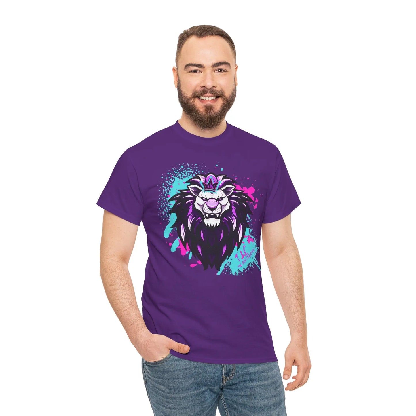 A smiling man with a beard is wearing The Lyon T-shirt, a purple garment featuring a colorful, stylized lion design. Made from 100% cotton using ethical production methods, he poses with one hand in his jeans pocket against a plain white background.