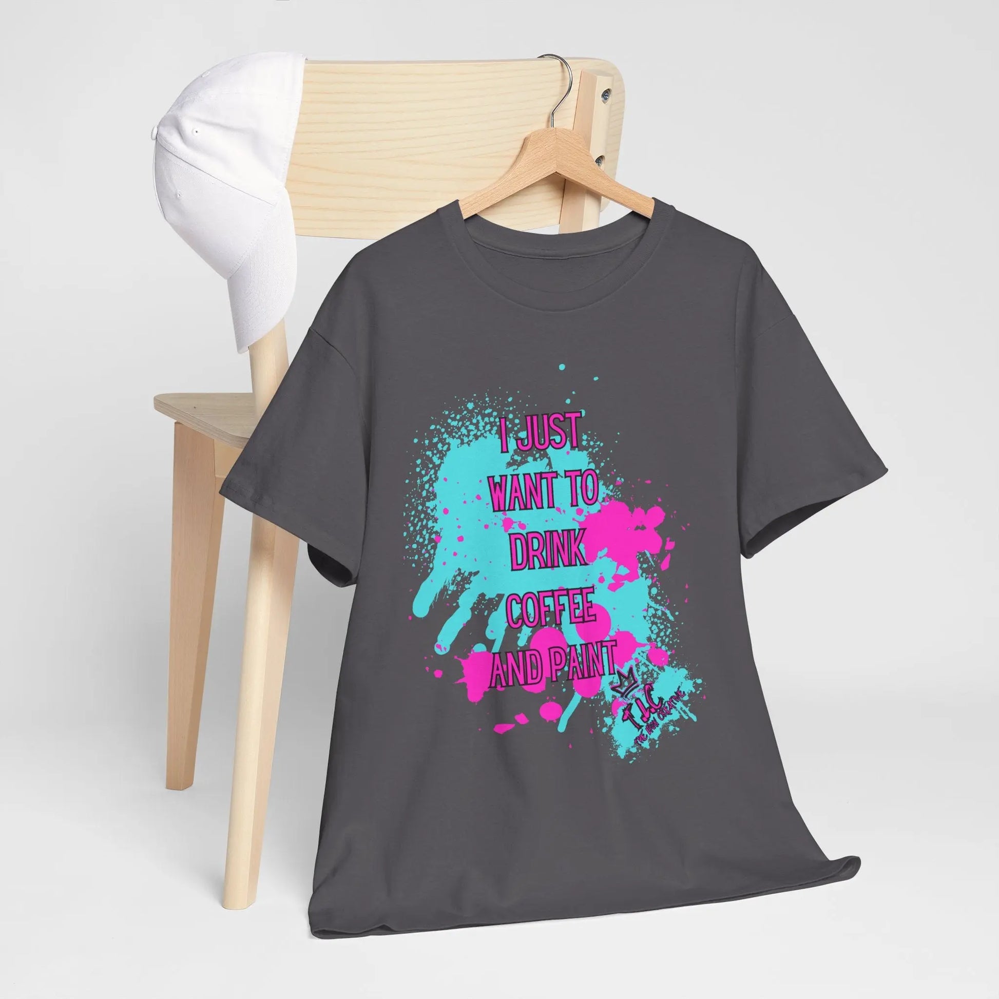 The "I Just Want to Drink Coffee" T-shirt, made from 100% cotton and featuring a dark gray base adorned with colorful paint splatters, hangs on a wooden chair. The text on the shirt reads, "I just want to drink coffee and paint." A white baseball cap is placed on the back of the chair.