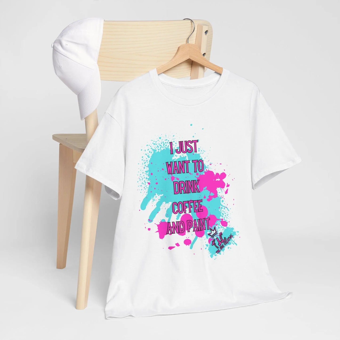 The "I Just want to Drink Coffee" T-shirt, made from 100% cotton and featuring colorful splashes with the phrase "I just want to drink coffee and paint," is draped over a chair with a white baseball cap perched on top. The classic fit tee is showcased against a plain background.