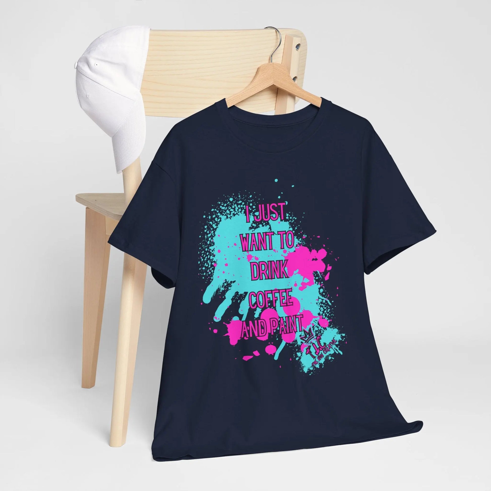 A black "I Just want to Drink Coffee" T-shirt, crafted from 100% cotton, drapes over a wooden chair, topped with a white cap. The T-shirt features vibrant paint splashes along with the text, "I just want to drink coffee and print.