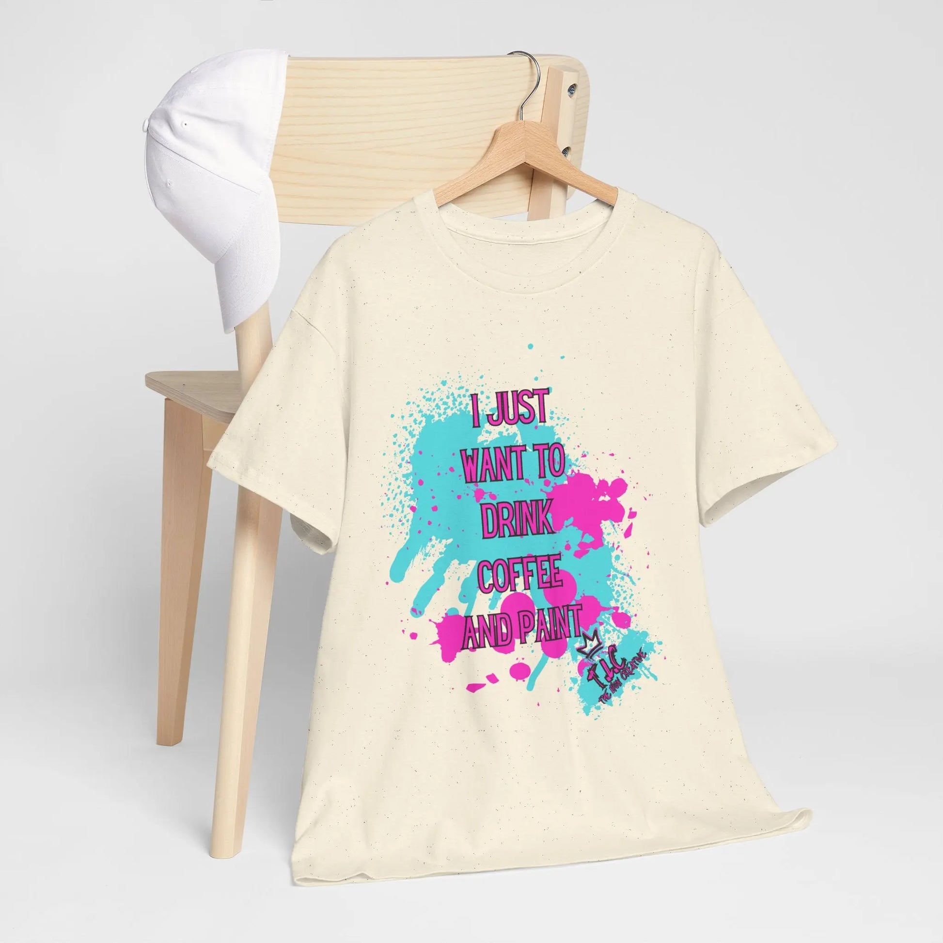 An "I Just Want to Drink Coffee T-shirt," crafted from 100% cotton in beige with vibrant paint splashes and text, hangs on a chair. A white cap is perched on top, rounding out the classic fit ensemble.