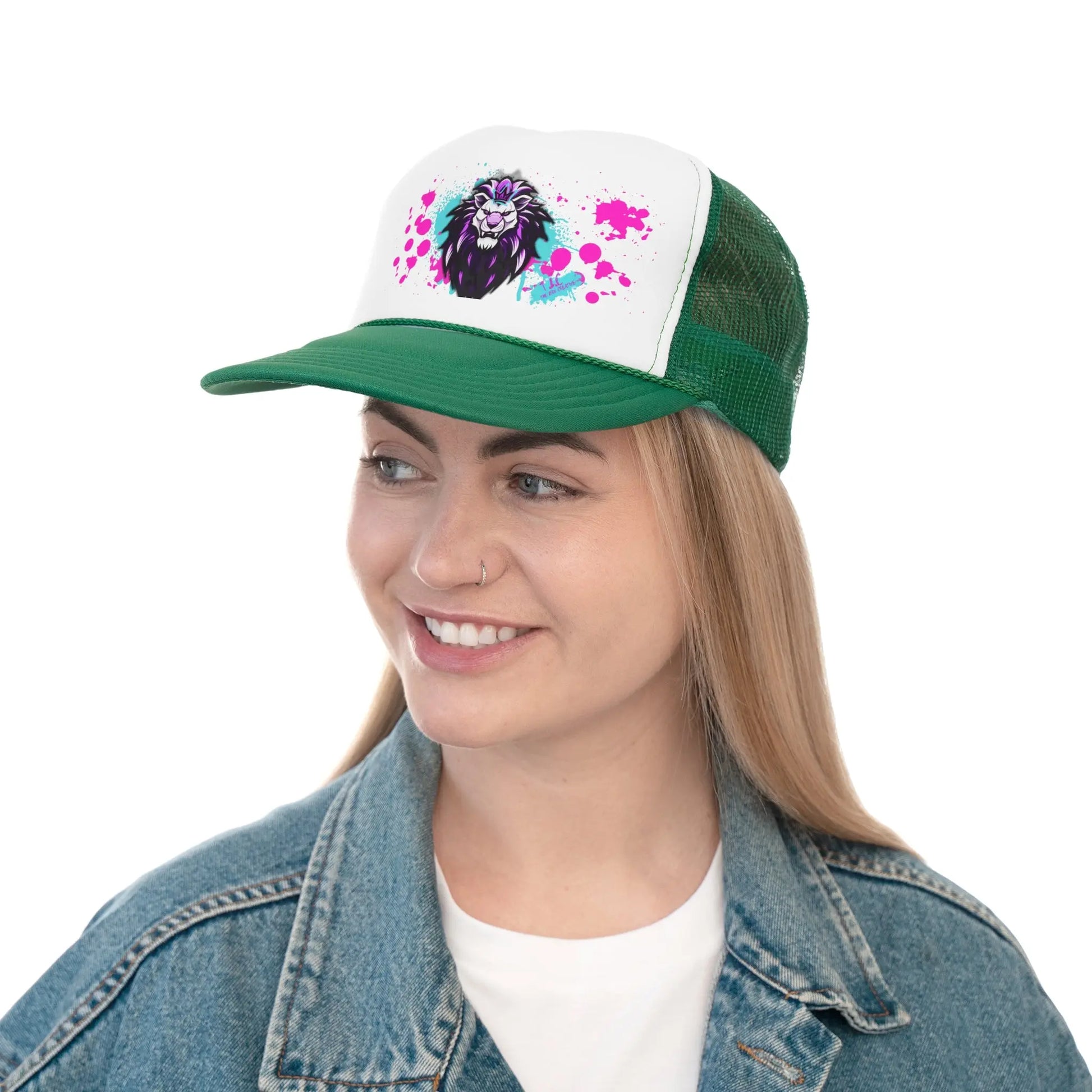 A person with long hair, wearing a denim jacket and a green and white Trucker Cap featuring a vibrant lion graphic with colorful splashes, is smiling while looking to the side. The cap has a polyester foam front and an adjustable snap closure, with the lion graphic primarily including black, pink, and blue colors.