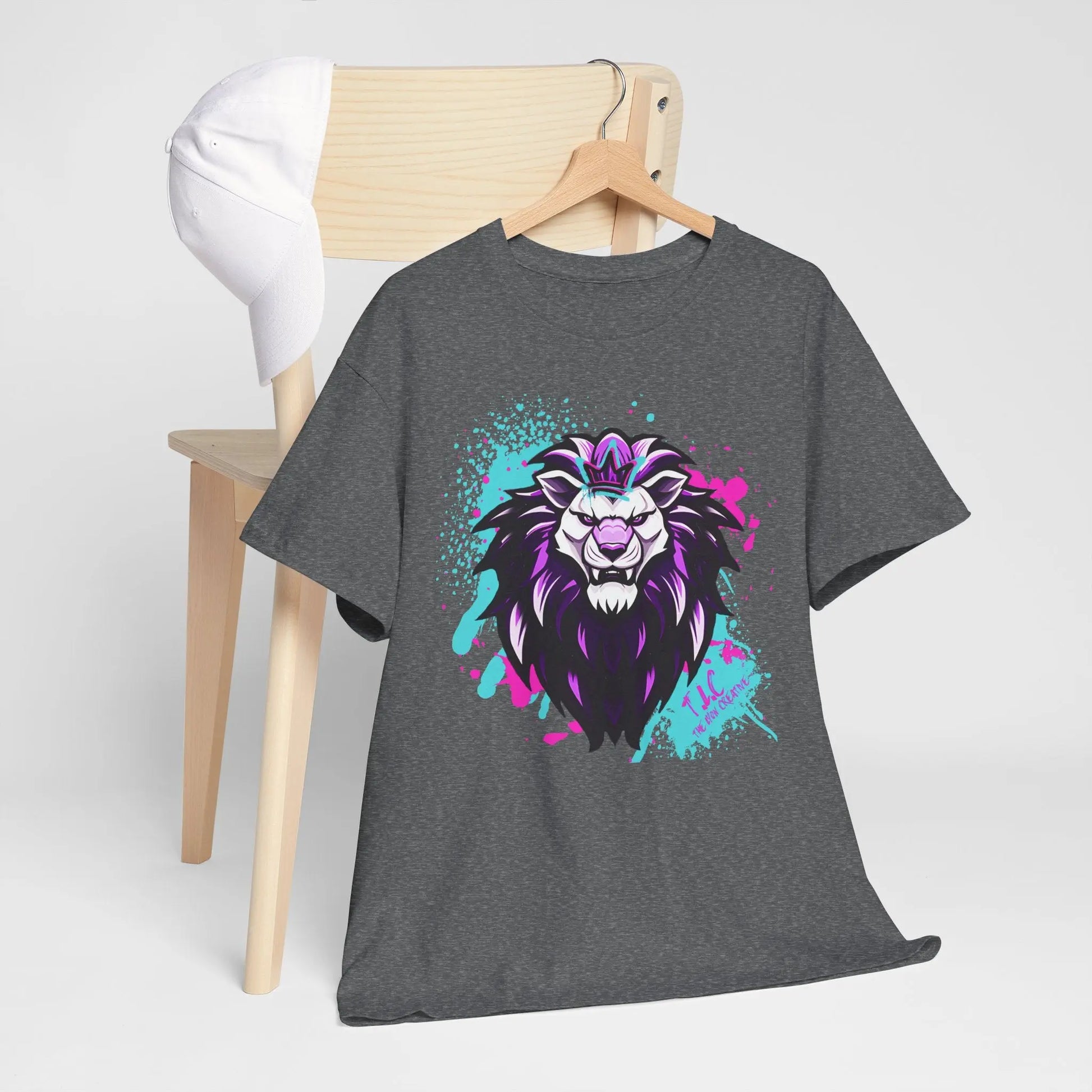 A gray T-shirt, known as The Lyon T-shirt, crafted from 100% cotton with a classic fit, showcases a colorful stylized lion graphic and is draped over a wooden chair. On the chair’s back sits a white cap. The design features purple and blue accents with splatters on a plain background, all produced sustainably.