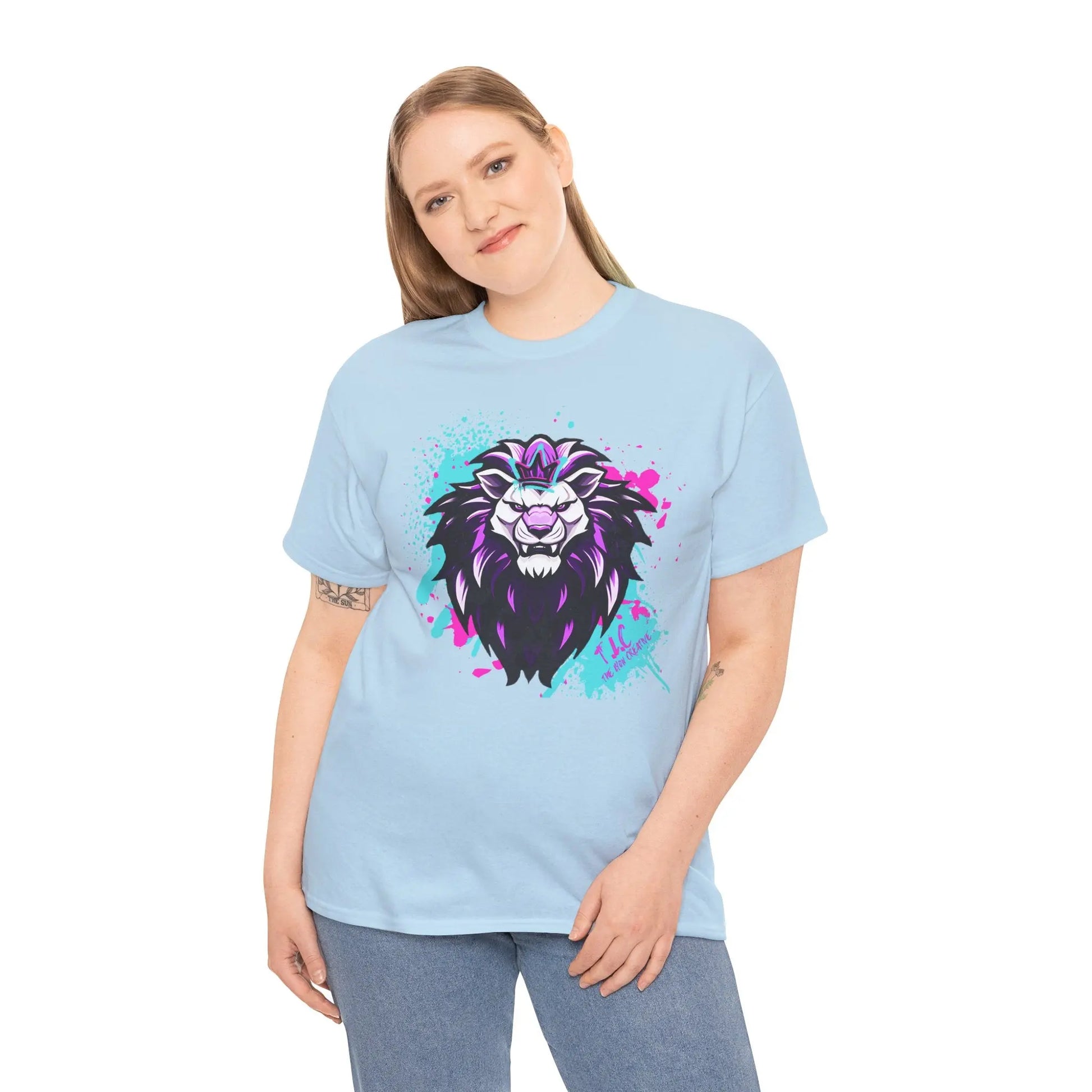 A person with long hair stands smiling, wearing The Lyon T-shirt, which is crafted from 100% cotton. It features a stylized lion with purple accents and offers a classic fit. The light blue background enhances the shirt's vibrant design while highlighting its sustainable choice.