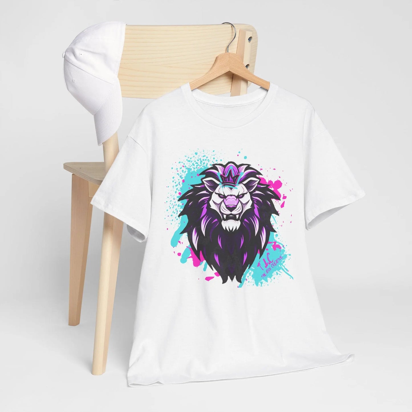 A classic fit, 100% cotton white t-shirt, known as The Lyon T-shirt, with a colorful lion design featuring purple and blue splatter accents on a pristine background, hangs on a wooden chair. A white baseball cap rests over the chair's back.