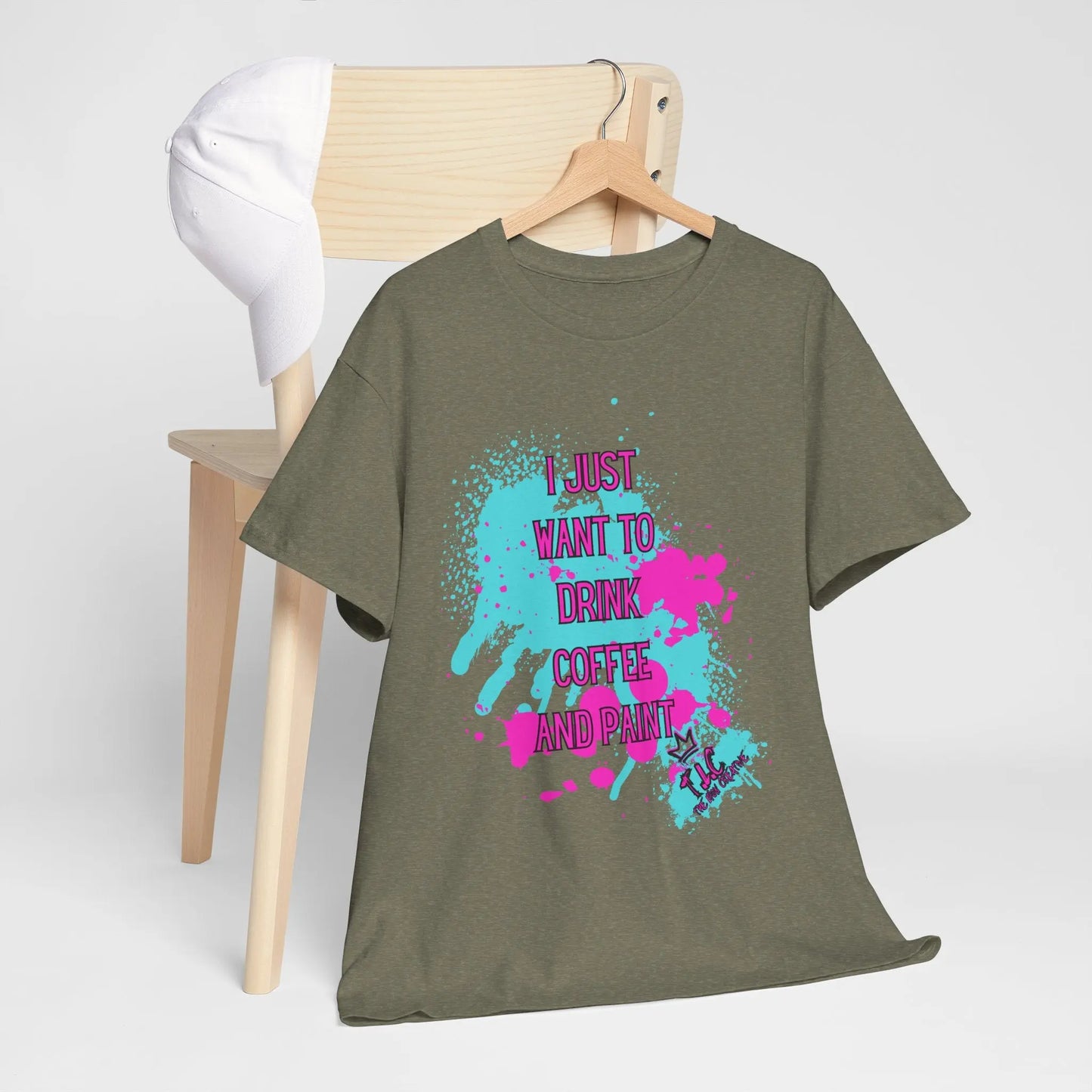 The "I Just Want to Drink Coffee" t-shirt, a classic fit in khaki green, is crafted from 100% cotton under the US Cotton Trust Protocol. It showcases a vibrant splash design featuring the phrase "I just want to drink coffee and paint." This shirt is displayed draped over a wooden chair, accompanied by a white baseball cap on the backrest against a plain white background.