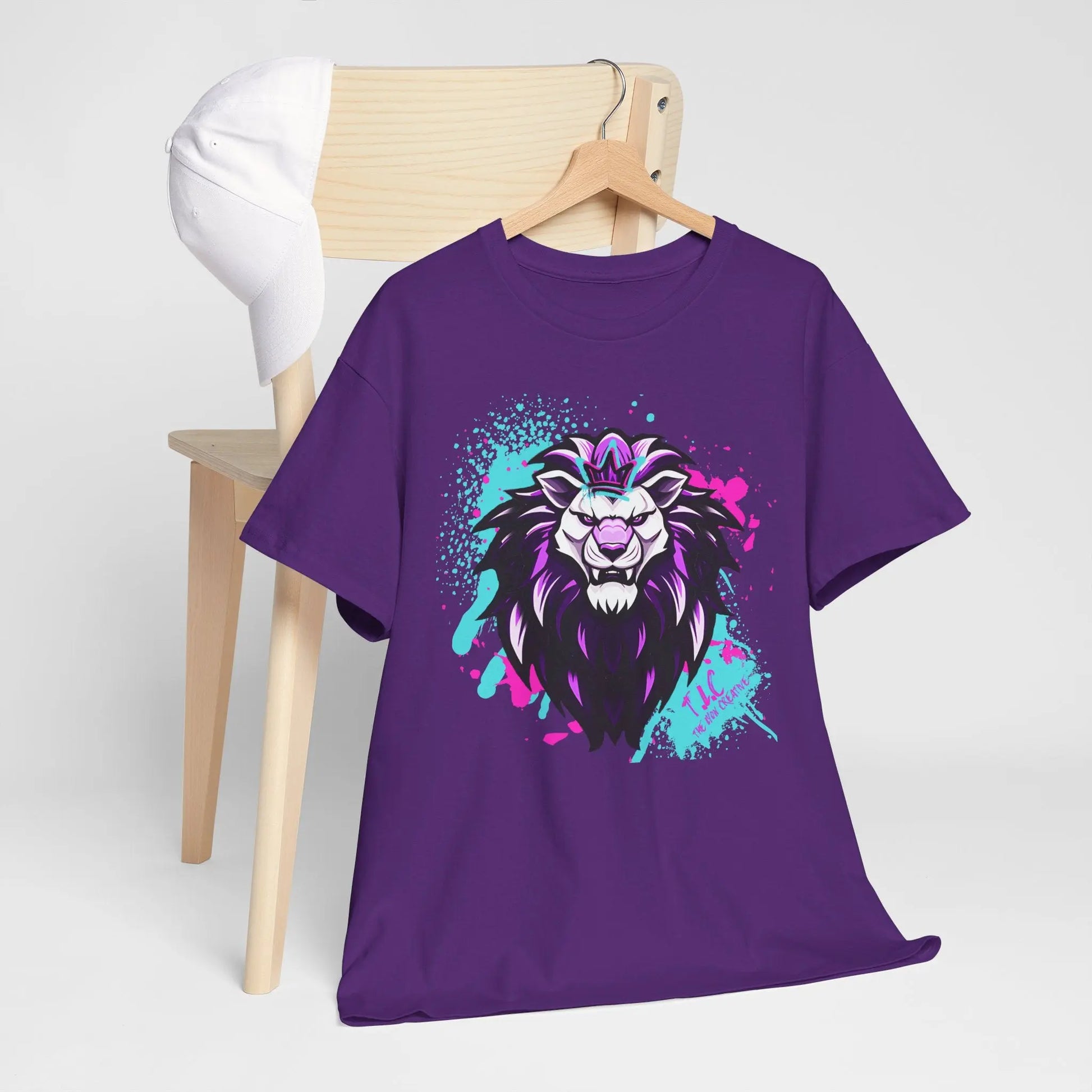 The Lyon T-shirt, made from 100% cotton and featuring a colorful, stylized lion graphic in purple, hangs on a wooden chair. A classic fit white baseball cap is placed on the chair's back. The plain white background accentuates the shirt's vibrant design and sustainable style.