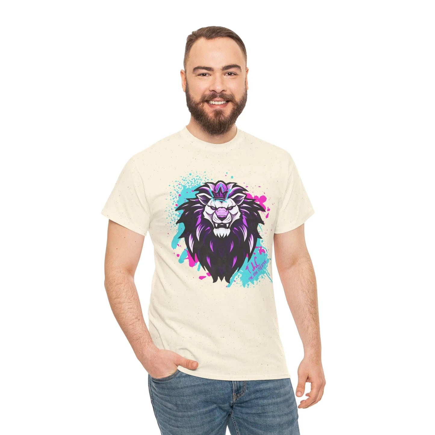 A man with a beard smiles while wearing The Lyon T-shirt, crafted from 100% cotton. The cream-colored shirt features a vibrant lion's head with a purple mane and colorful splatters. Ethically produced, he poses against a plain white background, one hand in his pocket.