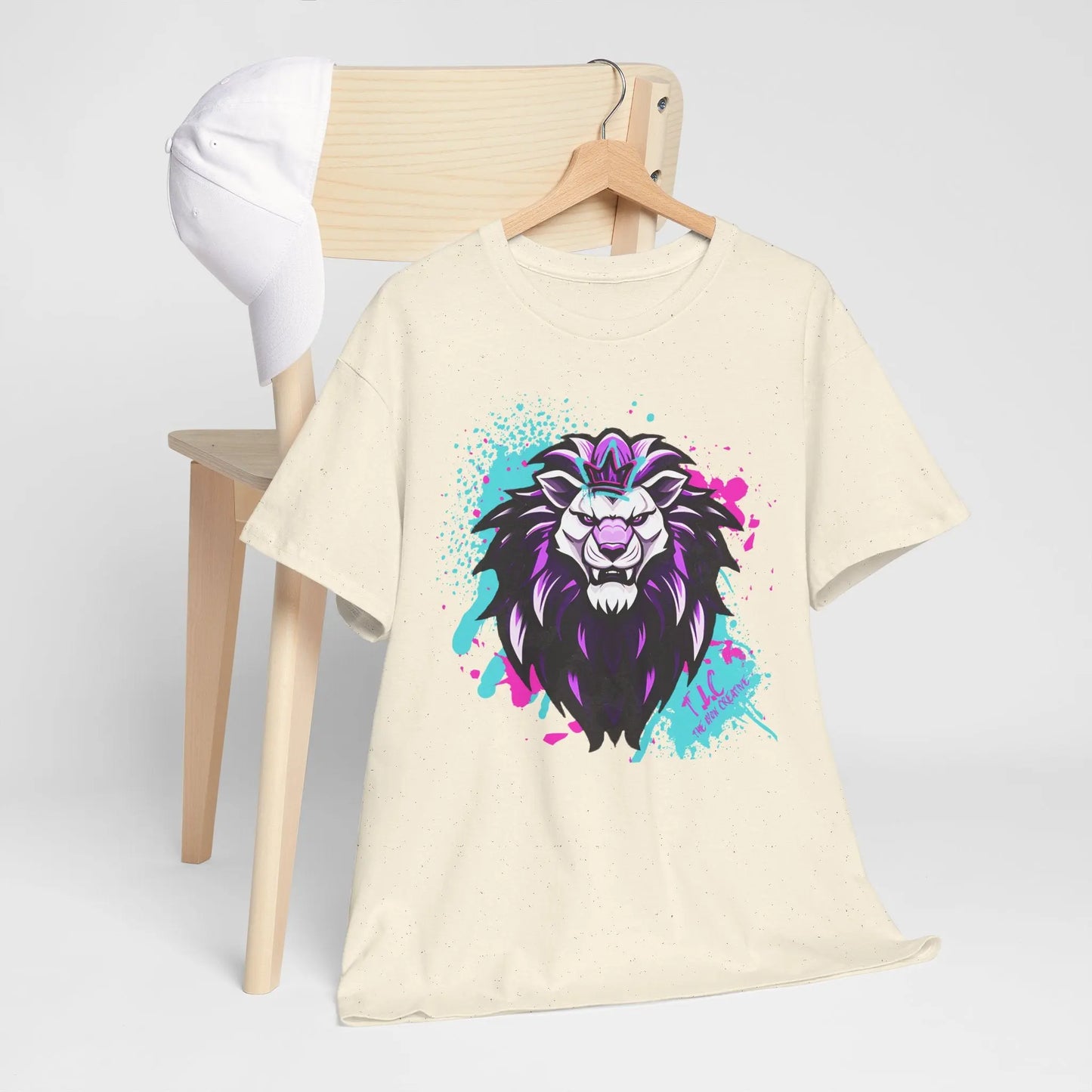 The wooden chair is adorned with The Lyon T-shirt, a beige classic fit made from 100% cotton, showcasing a vibrant lion graphic in purple and blue hues. A white cap is perched on the chair's backrest against a minimalistic plain background.