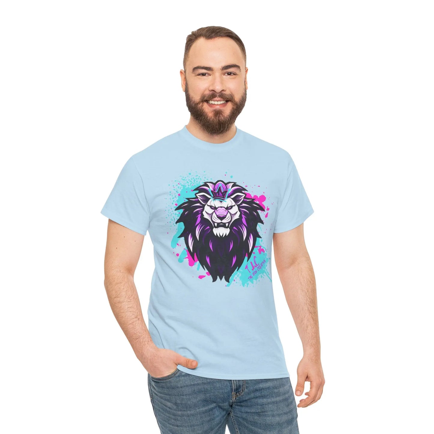 A man with a beard smiles while wearing The Lyon T-shirt, a light blue classic fit made from 100% cotton. It features a colorful lion graphic, with the lion sporting a purple mane accented by splashes of pink and teal. His relaxed style is complemented as he stands against a plain white background.