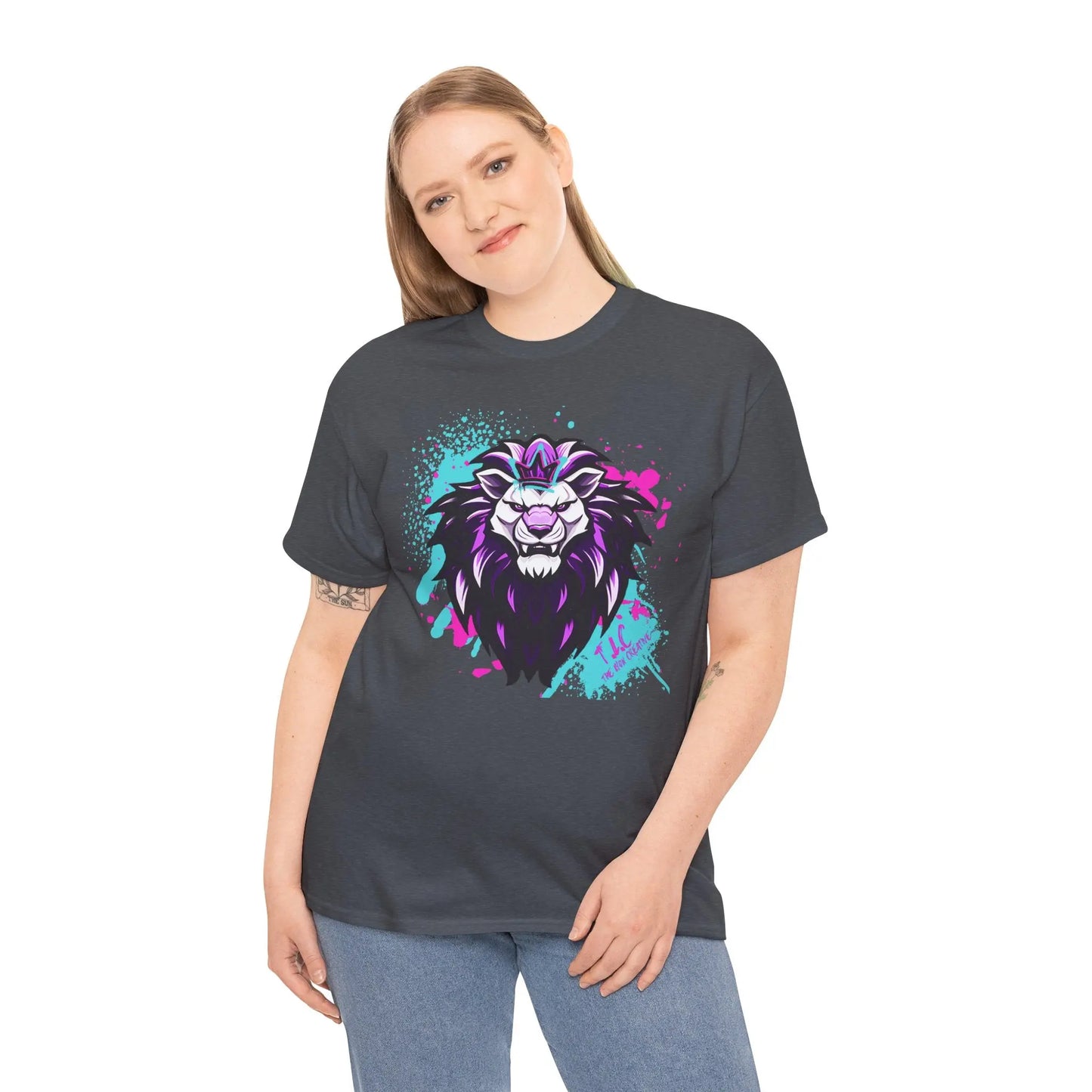 A person with long hair stands smiling, wearing The Lyon T-shirt in a classic fit dark gray, crafted from 100% cotton. This shirt features a vibrant lion graphic with purple and blue accents. Their forearms adorned with tattoos, they pair it stylishly with light blue jeans against a plain white background.