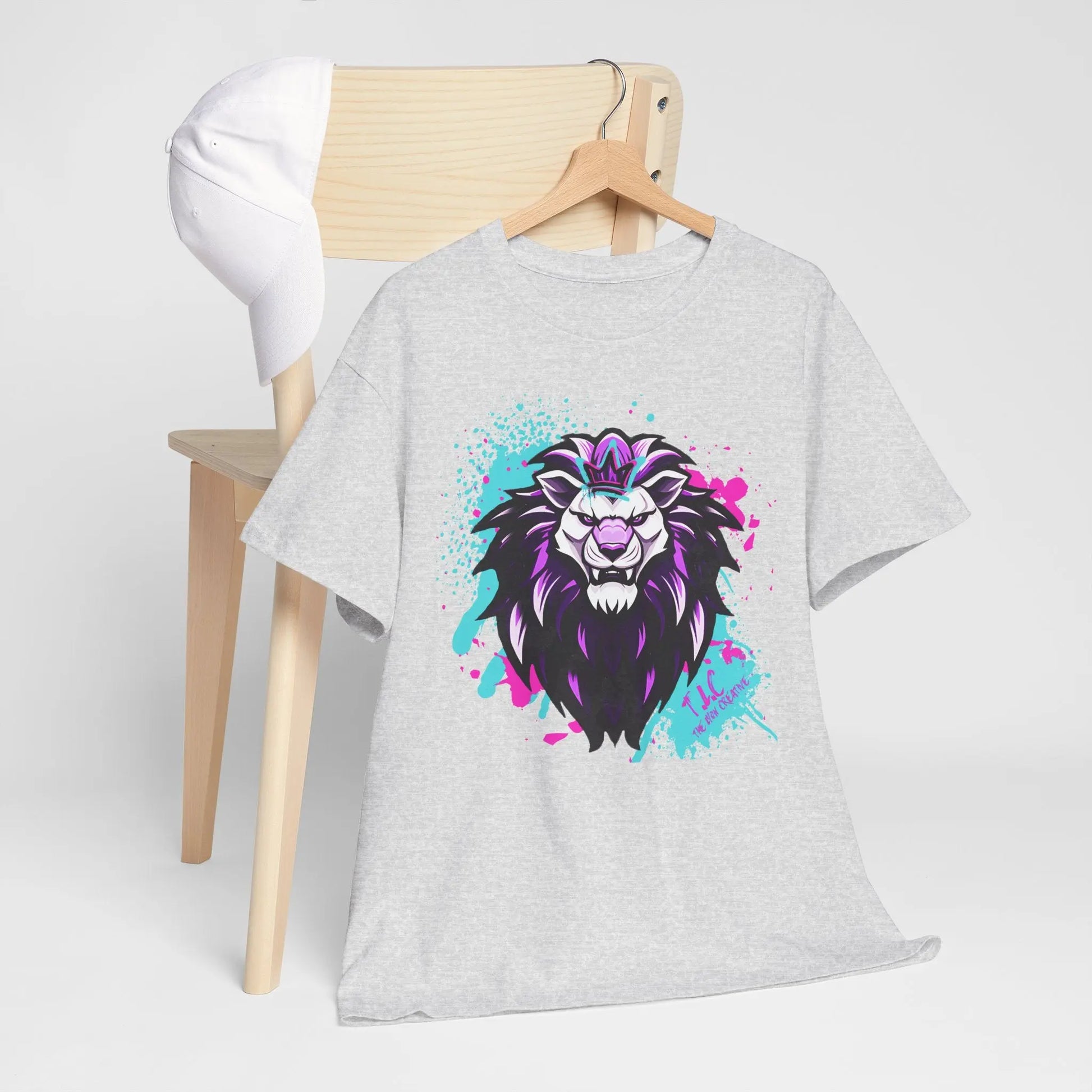 The Lyon T-shirt, crafted from 100% cotton and showcasing a classic fit with a vibrant lion graphic, is elegantly draped over a wooden chair. A white baseball cap sits on the backrest, suggesting eco-friendly production values.