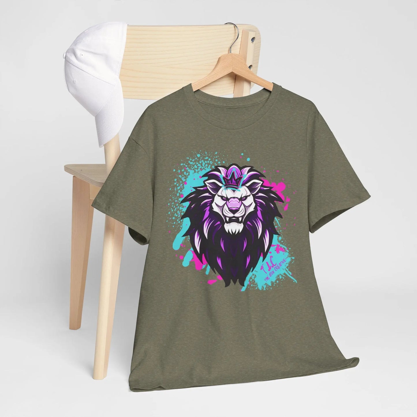 A green Lyon T-shirt, featuring a vibrant artistic lion face design and made from 100% cotton for a classic fit, hangs on a chair. A white baseball cap rests on the chair's backrest against a plain background.