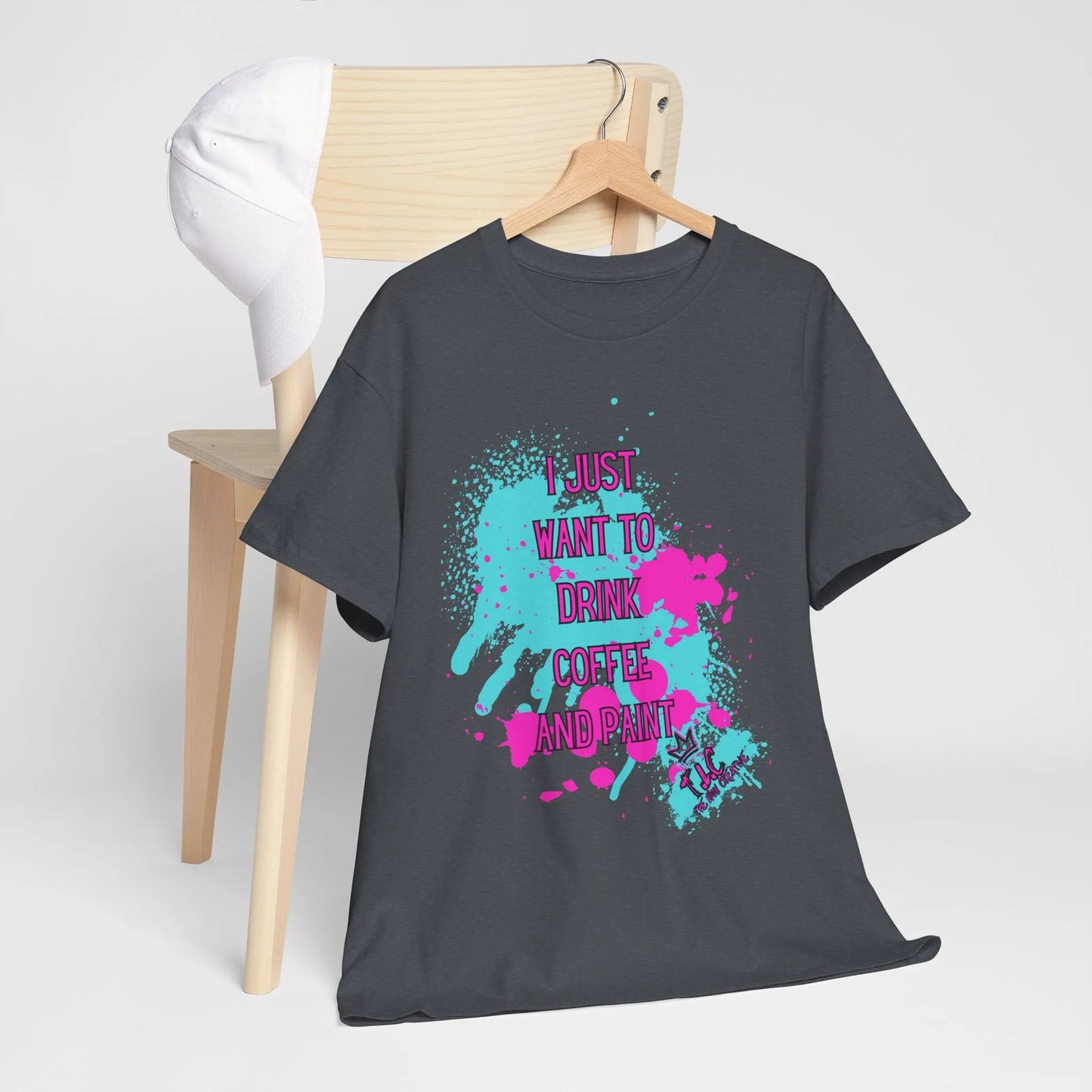 A "I Just Want to Drink Coffee" T-shirt made from 100% cotton in gray, featuring a classic fit, colorful paint splatters, and the phrase "I just want to drink coffee and paint," is draped over a wooden chair alongside a cap.