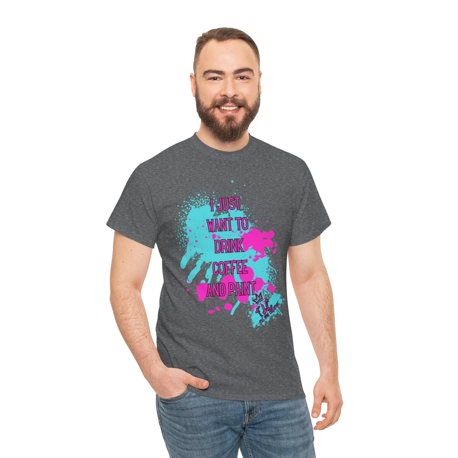 A man with a beard is wearing the "I Just Want to Drink Coffee" T-shirt, an ethically made gray cotton shirt that showcases a colorful splatter design and the text, "I just want to drink coffee and print." He stands smiling against a white background, with one hand casually in his jeans pocket.