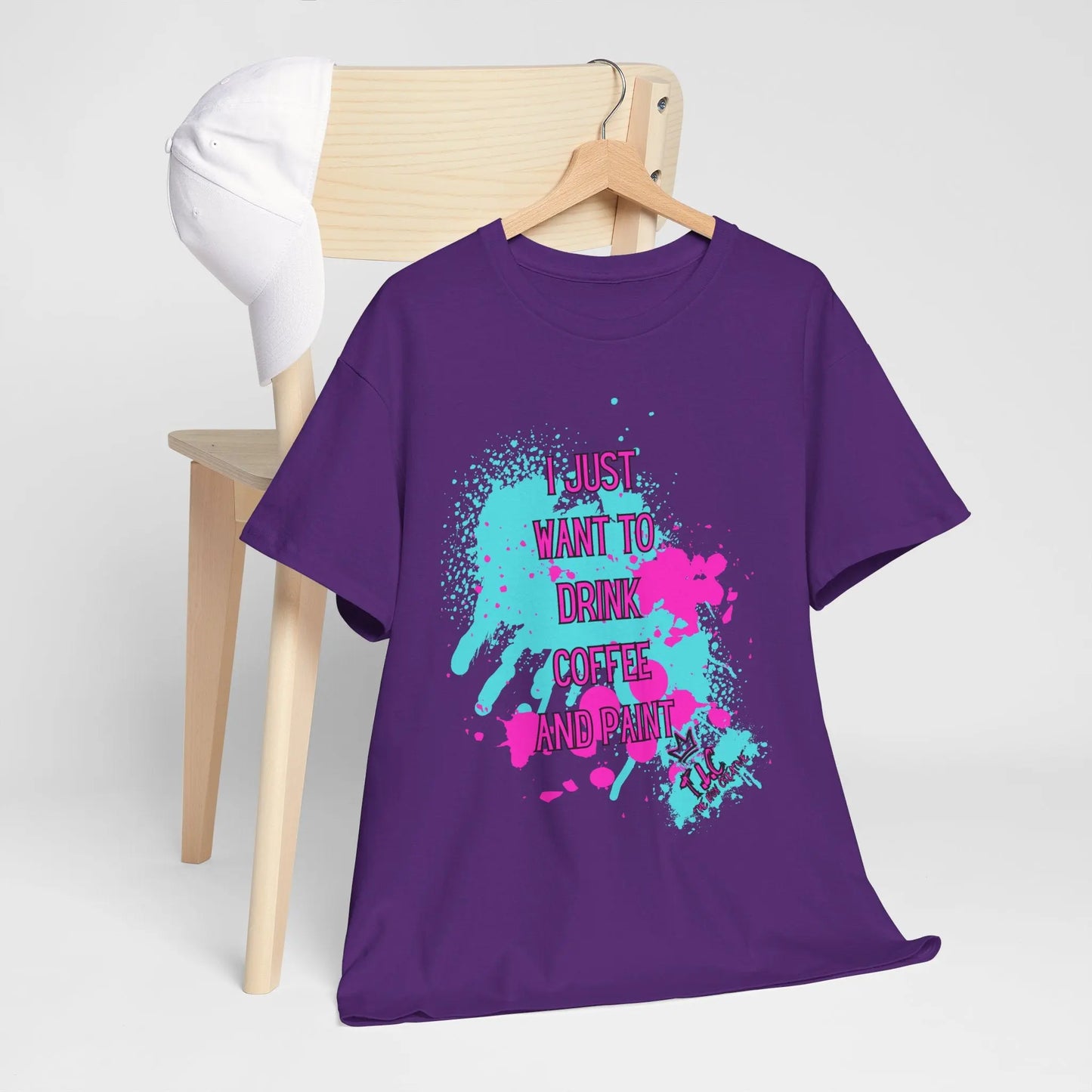 The "I Just Want to Drink Coffee T-shirt," featuring a sustainable, classic fit design and bold-text message, hangs on a wooden chair. This purple T-shirt is crafted from 100% cotton and pairs well with a white baseball cap draped over the chair's backrest.