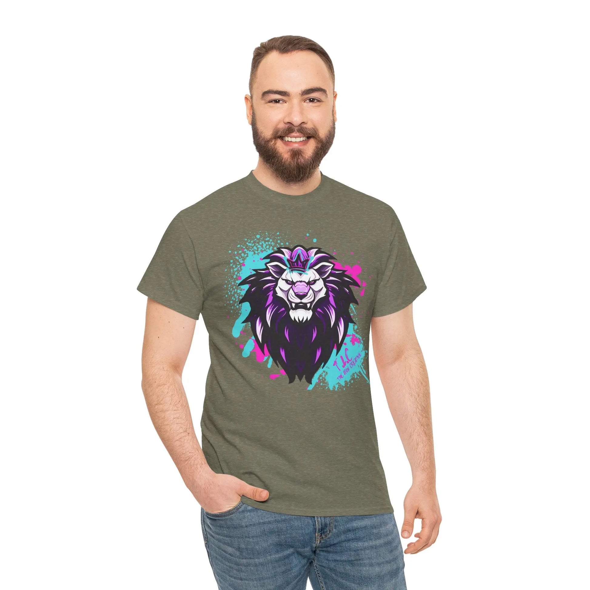 A man with a beard is smiling and wearing The Lyon T-shirt, a green, 100% cotton shirt featuring a colorful lion graphic. His right hand is in his pocket against a plain white background.