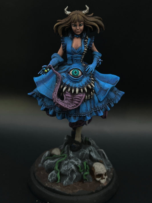 This 3D printed miniature, Mary The Mimic Girl - Painted Miniature, features a woman in a blue dress with an eye and teeth design on the front, perfect for your Dungeons and Dragons campaign. She has small horns on her head, stands on rocky terrain with skulls and greenery, and holds a small yellow object in her left hand.