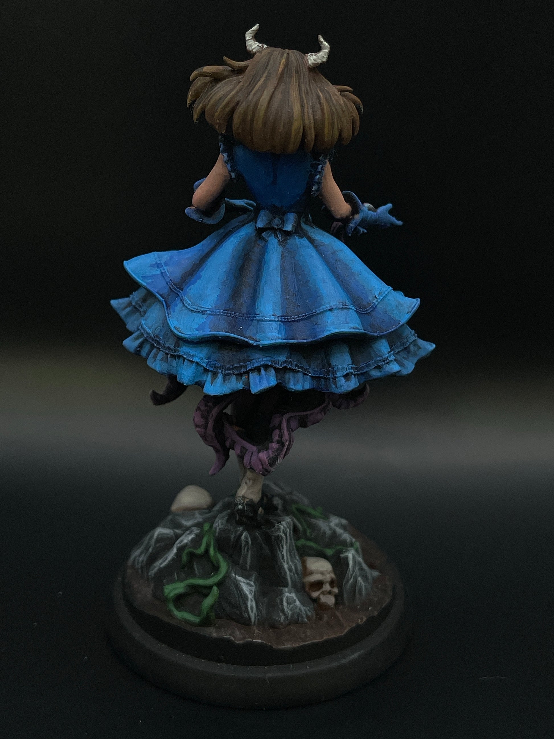 A 3D-printed miniature of Mary The Mimic Girl - Painted Miniature wearing a blue dress, brown gloves, and a headpiece with small horns stands on a rocky terrain base adorned with vines and skulls. The figure is facing away, showcasing the dress's flowing and layered design—perfect for any Dungeons and Dragons campaign.